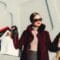 Woman with burgundy coat and pink sweater wearing dark sunglasses, holding multiple shopping bags on both sides outdoors.