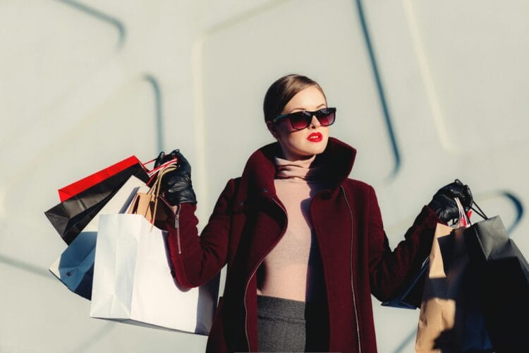 Woman with burgundy coat and pink sweater wearing dark sunglasses, holding multiple shopping bags on both sides outdoors.