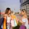 Two women chatting outside a modern building, each carrying multiple colorful shopping bags in bright daylight.